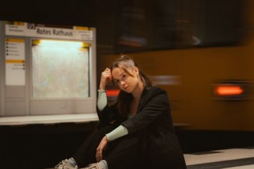 A young woman in casual attire seated at a Berlin subway station with trains in motion.